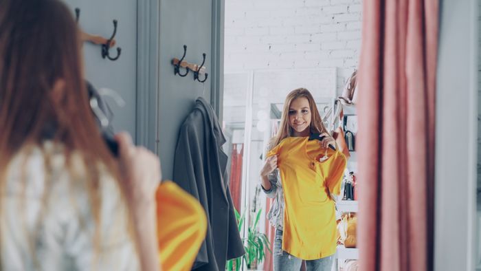 A woman in a fitting room holding a marigold top up to the mirror